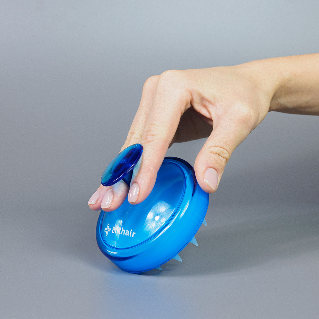 Eine Hand hält die Dr. Balwi Massagebürste von shop.elithairtransplant mit einem blauen runden Griff und kurzen, flexiblen Borsten – ideal für Kopfmassage und Kopfhautpflege – vor einem schlichten grauen Hintergrund.